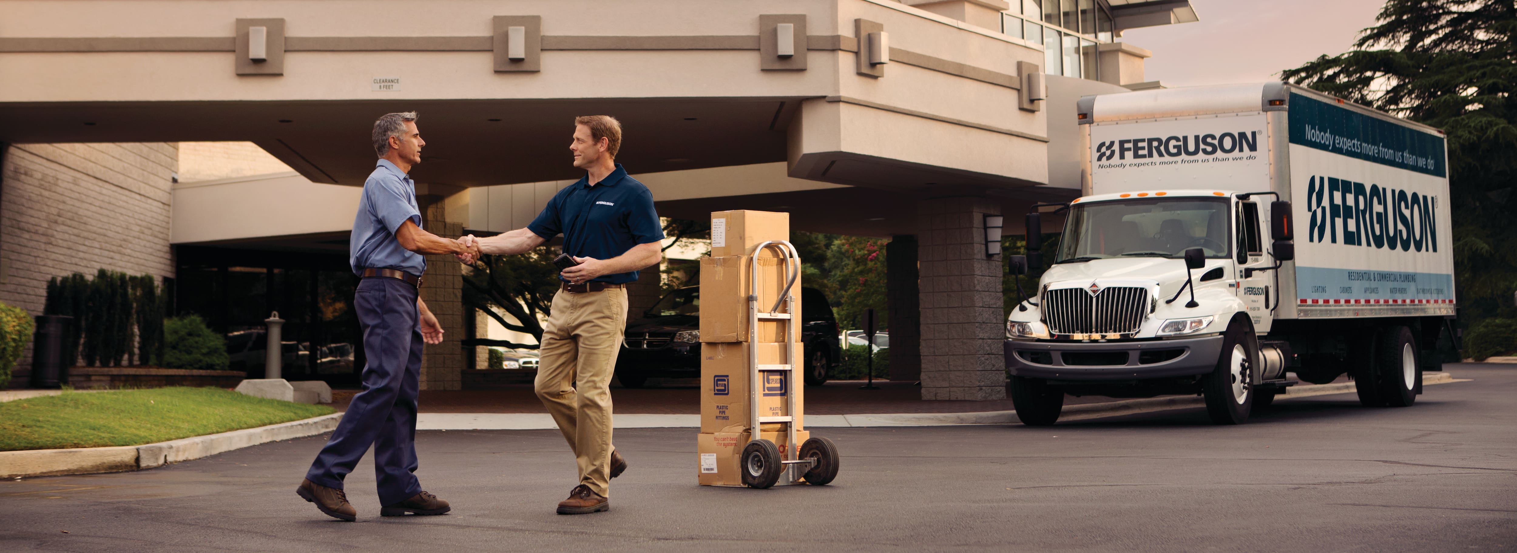 A Ferguson associate shakes hands with a hospitality manager in front of a hotel. A Ferguson truck and a dolly with Ferguson packages are behind the pair.