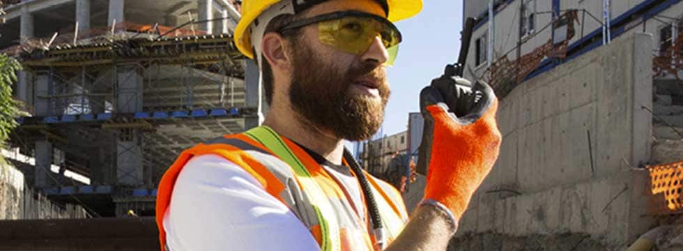 A worker wearing reflective PPE speaks into a radio on a construction jobsite.