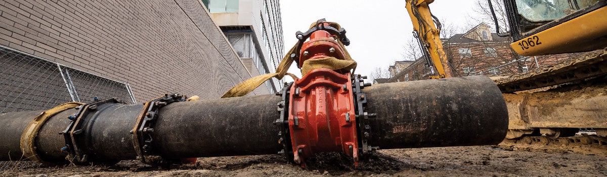 A close-up of an underground utility pipe lying in an excavated alley space, with an excavator parked on the right side.
