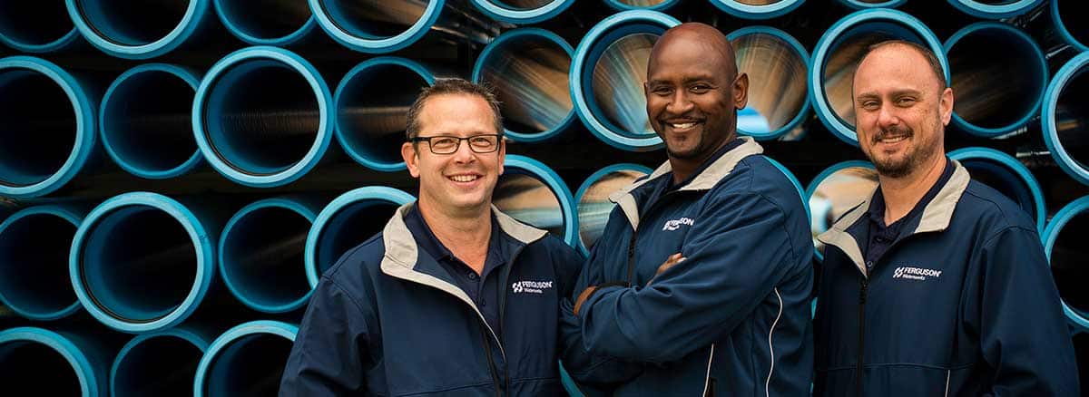 Two smiling Ferguson associates stand in front of a stack of large pipe, with sunlight streaming through the ends.