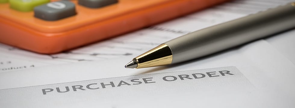 A close-up view of a purchase order with a silver pen lying across it and an orange calculator partially in sight.
