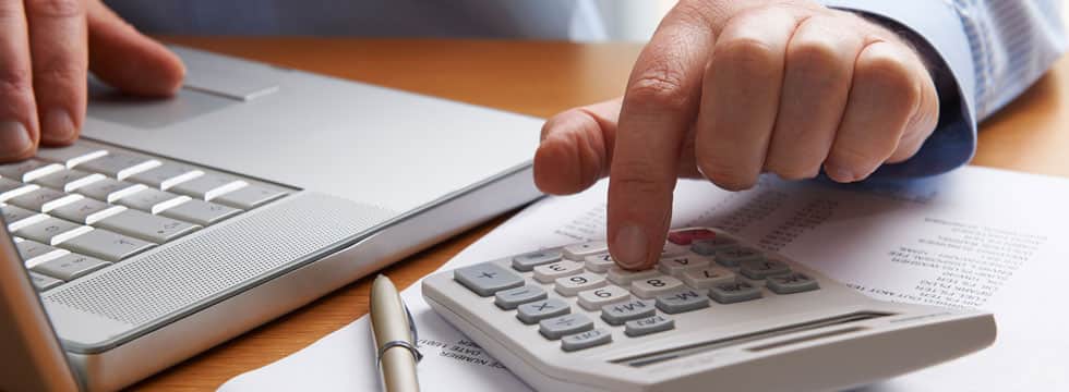 A person's hand presses numbers on a calculator, with a pen and paper next to it on a desk.