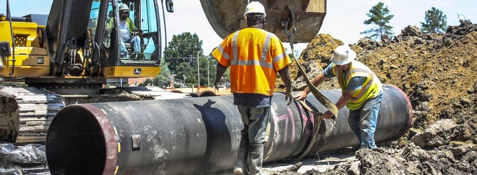 Two workers in hard hats remove a ratchet strap from a large underground utility pipe, which a driver in a front-end loader has just set down on the jobsite.