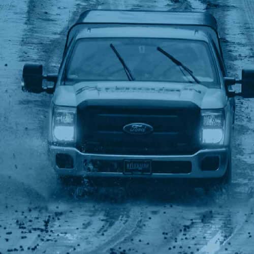 A Ferguson work truck drives on a wet road during a rainstorm, with headlights and windshield wipers on.