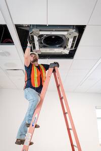 An HVAC contractor wearing an orange safety vest stands on a ladder and inspects the exhaust fan of a heating and cooling system.