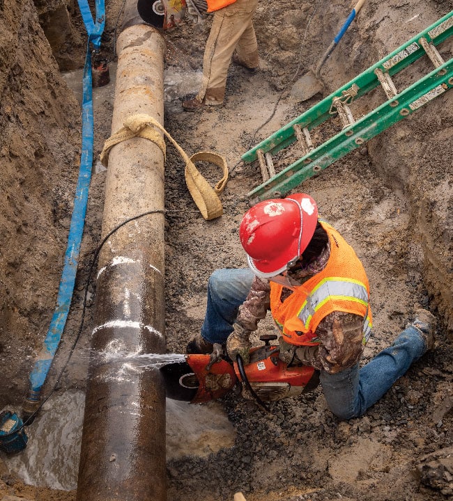A worker in a trench cuts through a large underground utility pipe with a circular saw and water attachment.