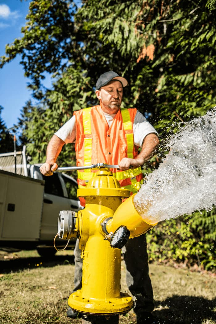 A worker wearing a reflective vest wrenches open a fire hydrant while water flows out.