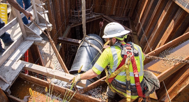 A worker wearing a white hard hat, yellow reflective vest and fall protection gear watches from above as a large utility pipe is lowered into a shaft.
