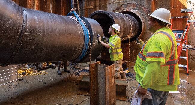 Two workers wearing white hard hats and yellow reflective vests guide the connection of two large underground utility pipes.