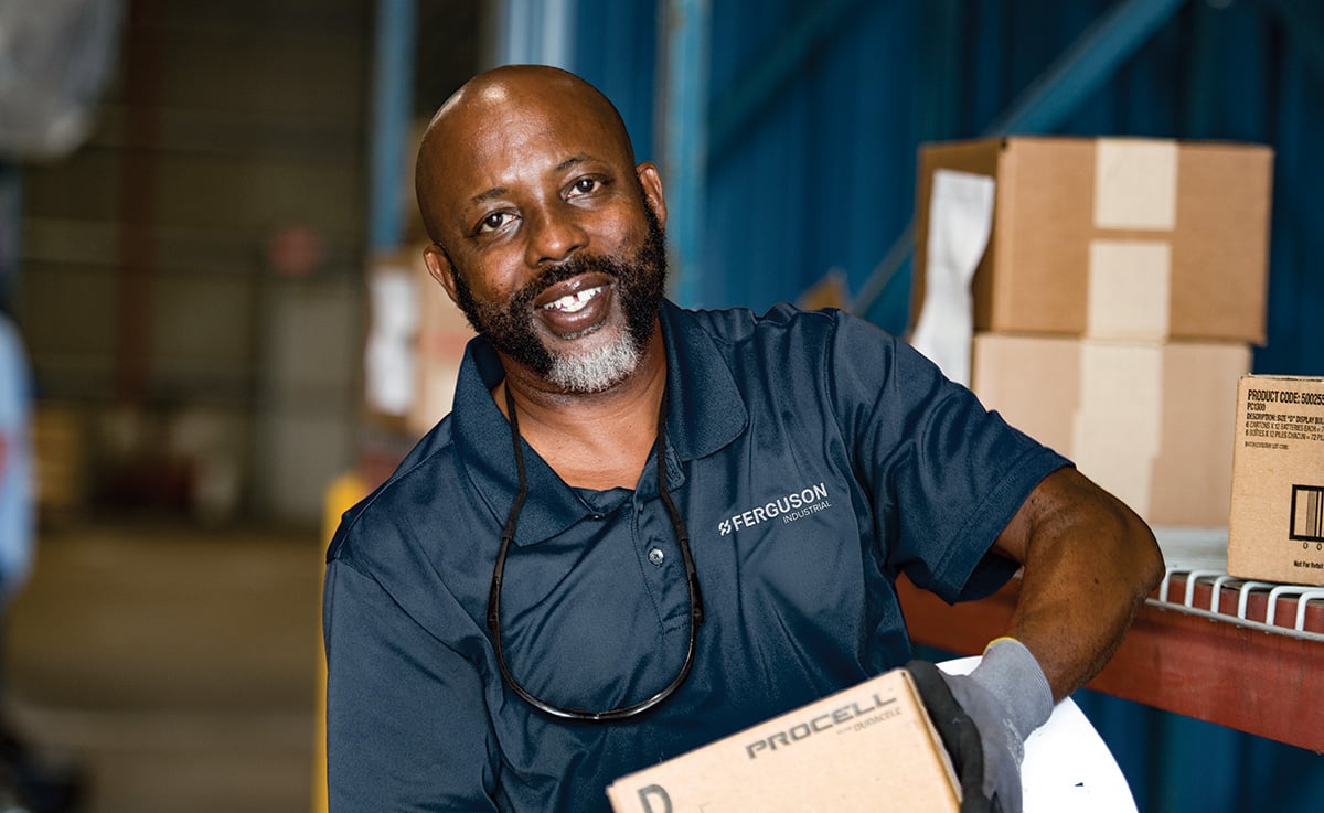 A Ferguson associate in the warehouse holds a box for shipping in front of a shelf of packaged boxes.