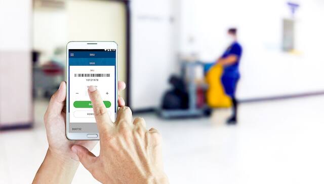 A hand points to a product barcode on ferguson.com on a smartphone in an institutional building hallway. A custodial engineer with cleaning and hygiene supplies is blurred in the background.
