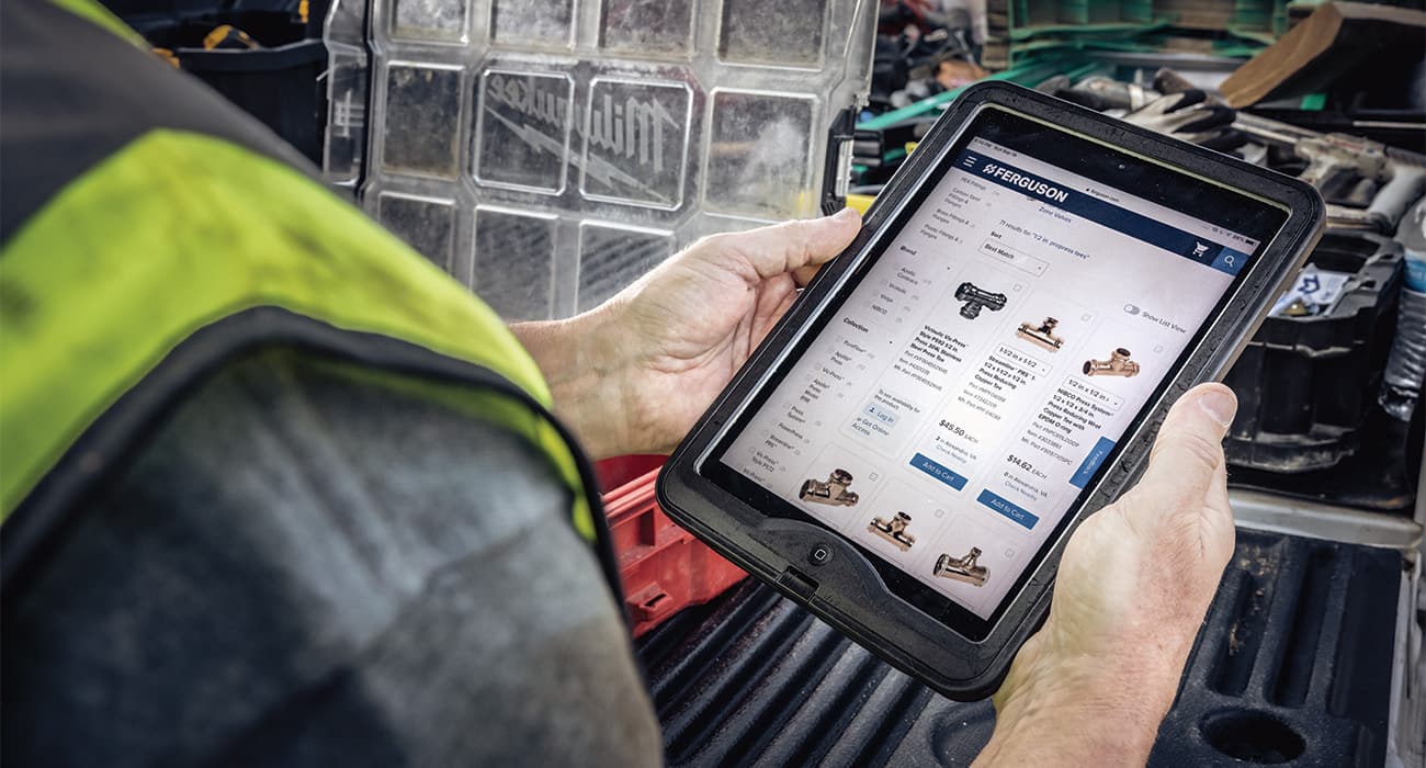 A contractor wearing a reflective vest holds a tablet open to a Ferguson products page.