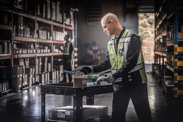 A Ferguson associate wearing a reflective vest examines an order of pipe brackets on a wheeled utility cart in a warehouse.