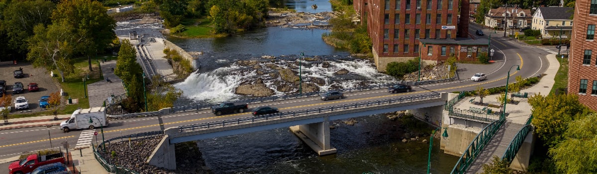 Vehicles travel on a bridge over a splashing river. Buildings and homes are on either side of the bridge.