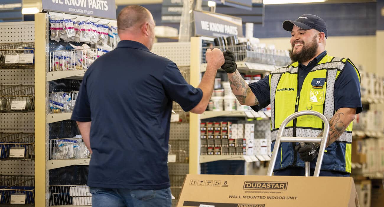 A Ferguson associate at a counter location pushes a dolly with a wall-mounted indoor HVAC unit and fist bumps a contractor.