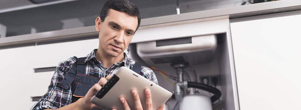 A plumbing professional stares intently at a tablet in a kitchen, the under-sink cabinet open behind him.