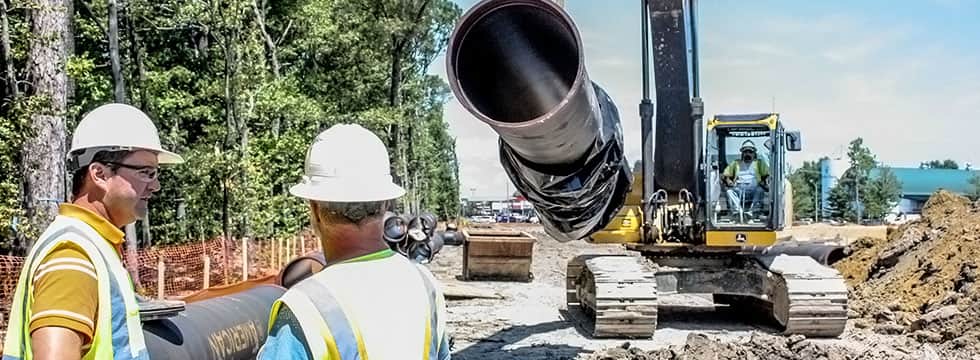 A waterworks utility contractor operates a dozer carrying a large underground pipe, and two other workers talk in the foreground.