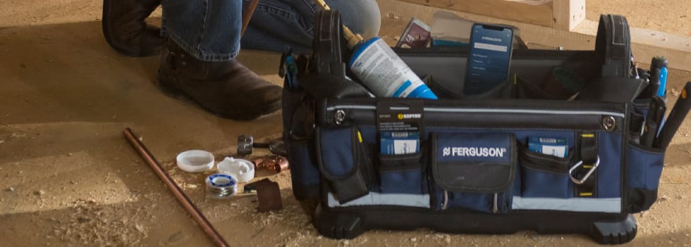 On a new construction site, a contractor kneels behind a Ferguson work bag, with various supplies tucked into the pockets, and the Ferguson app open on a mobile phone.
