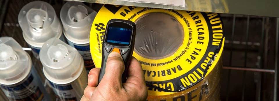 A hand holds a scanning device over a stack of barricade tape in a warehouse.